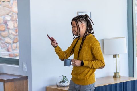 African american man standing in modern living room holding smartphone and mug