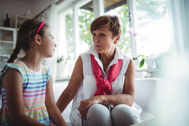 Senior woman and her granddaughter sitting together at home, engaging in a heartfelt conversation. Ideal for use in family-oriented content, advertisements promoting family values, or articles about intergenerational relationships and bonding.