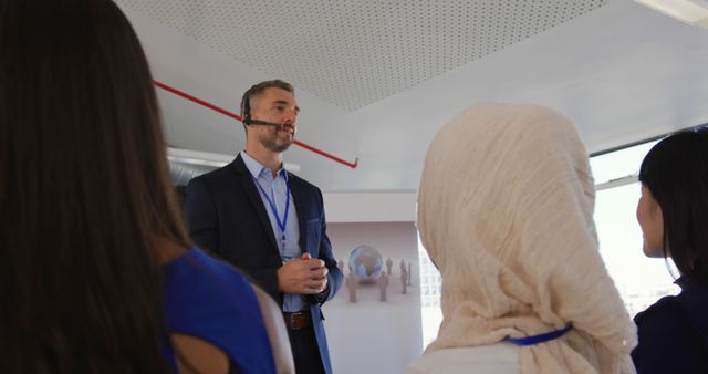 Business speaker wearing headset giving a presentation to a diverse team in a conference room. Ideal for use in articles and publications about business communication, team collaboration, and professional meetings. Highlights multicultural interactions in a corporate setting.