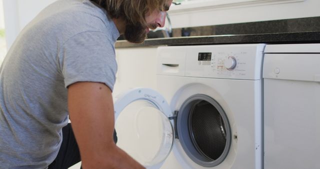 Man is loading clothes into a front loading washing machine in a modern home laundry room. Ideal for content related to home chores, modern household tips, cleaning, and lifestyle blogs. Can be used in advertisements for home appliances or laundry products.
