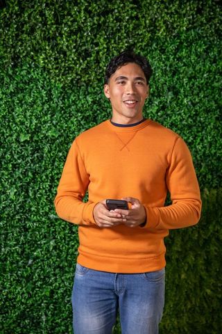 Young asian man wearing orange sweatshirt holding smartphone in front of green hedge