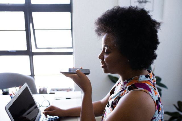 This image shows an African American woman using a smartphone and laptop in a home office setting. She appears to be engaging in online communication, possibly for remote work or staying connected during self-isolation. This image can be used for articles or advertisements related to remote work, technology, home office setups, or the impact of the coronavirus pandemic on daily life.