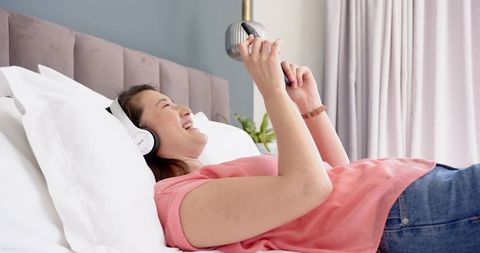 Asian Woman Relaxes with Smartphone and Headphones on Bed