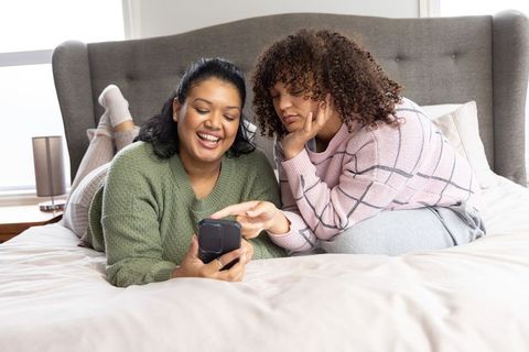 Indian and african american women reclining on bed sharing smartphone, laughing and smiling