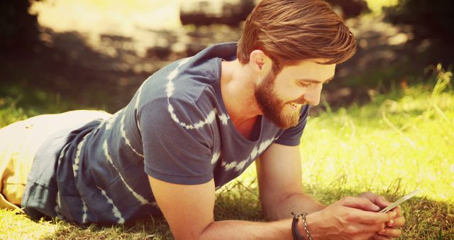 Young bearded man smiling while using smartphone, lying on grass in a casual setting. Perfect for illustrating concepts related to leisure, communication, and lifestyle. Useful for advertisements, blog posts, social media campaigns focusing on young adults, outdoor activities, and technology use.