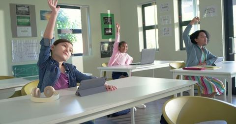 Elementary school girls raising hands while using tablets in bright modern classroom