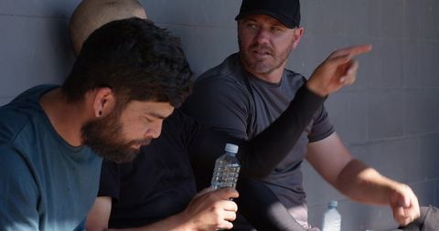 Sunlit baseball teammates relaxing in dugout pointing and hydrating between innings