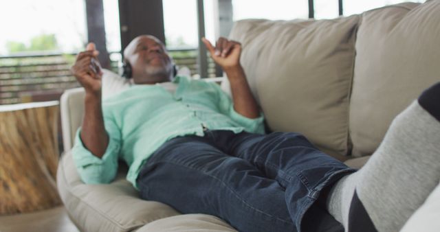 Elderly man is lounging on a comfortable couch, enjoying music through his headphones. He is wearing a green shirt and jeans, looking content and relaxed. This image is ideal for topics related to leisure activities, senior lifestyle, relaxation, or promotional materials for audio devices and music services.