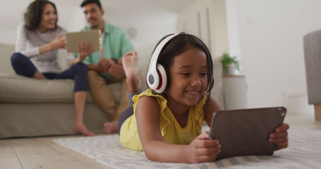 Young girl lying on the floor using a tablet, enjoying digital content while wearing headphones. Parents sit on sofa in the background, engaging with their own devices, creating a relaxed family environment. Ideal for illustrating modern family bonding, technology integration in daily life, or remote learning scenarios.
