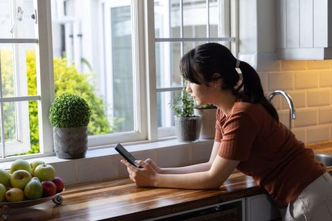 Woman leaning on kitchen counter checking smartphone by window with plants and fruit