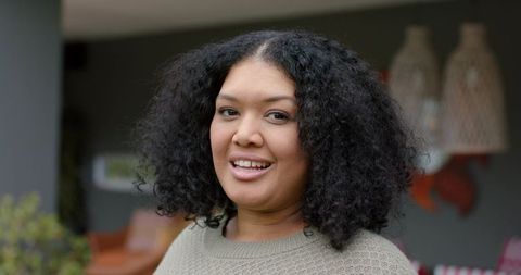 Smiling woman with curly hair wearing beige knit sweater on veranda with woven lamps