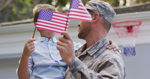 Depicting a heartfelt reunion between a military father and his son, both holding American flags. Useful for themes involving patriotism, familial bonds, military homecomings, celebrations such as Memorial Day and Independence Day, and promotional materials for veteran support services and military family events.