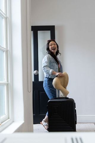 Asian woman leaving home with suitcase and straw hat smiling in bright entryway