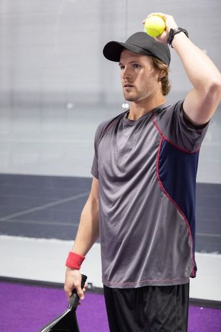 Focused male player serving with paddle and yellow ball on indoor purple court