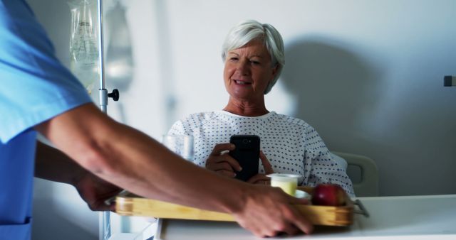 This stock image features a senior woman patient in a hospital room receiving a meal tray from a nurse. The patient is dressed in a hospital gown and appears to be smiling, suggesting a positive mood and supportive care environment. This image can be used in contexts related to healthcare, hospital care, patient recovery, elderly care, and medical services marketing.