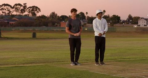 Standing diverse friends on cricket pitch at dusk holding cricket ball, casual athletes