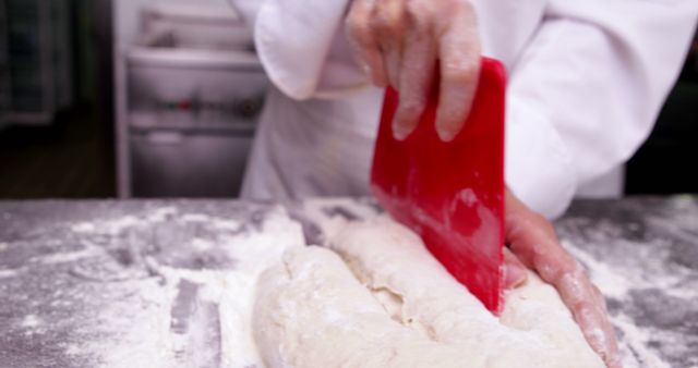 Baker is working with dough using a red scraper on a floured surface in a professional kitchen. Ideal for use in articles or websites about baking, artisanal bread making, pastry cooking classes, and professional culinary training.