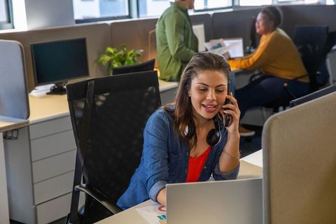 Woman in 30s speaking on smartphone while working on laptop in modern open office