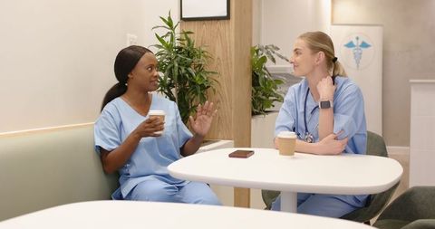 Healthcare colleagues chatting over coffee in clinic break room wearing scrubs and stethoscope