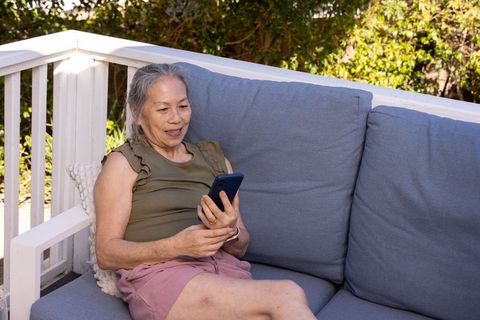 Senior asian woman relaxing on deck sofa using smartphone on sunny patio