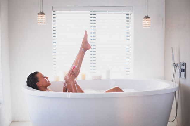This image depicts a biracial woman lying back in a modern bathtub, shaving her leg. The bathroom features a clean, minimalist design with white walls and blinds, and elegant hanging lights. This image is ideal for use in articles or advertisements related to self-care, personal grooming, home lifestyle, and quarantine activities. It can also be used in content promoting relaxation, hygiene products, or luxury bathroom fixtures.