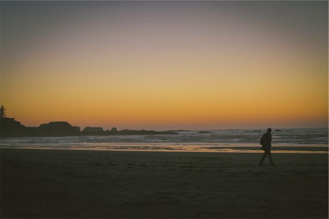 Person walking along beach as sun sets, creating tranquil and peaceful scene. Beautiful horizon with distant rocks enhancing natural beauty. Ideal for themes of relaxation, solitude, vacation, and nature. Suitable for blogs, travel websites, and relaxation content.