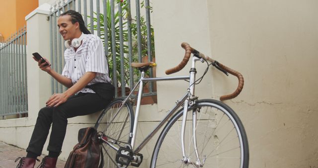 Urban cyclist taking a break while using a smartphone. Person is sitting on curb beside bicycle, dressed in casual attire with headphones around neck. Fits well with themes of mobile technology, modern transportation, and lifestyle. Ideal for promoting cycling gear, mobile apps, or urban living concepts.