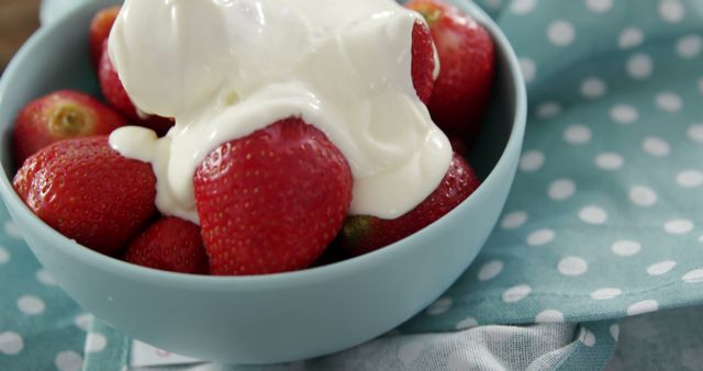 A bowl of fresh strawberries topped with a dollop of creamy yogurt sits on a table with a polka-dotted cloth. This delicious and healthy snack is a perfect combination of natural sweetness and creamy texture.