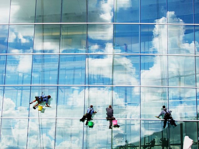 Professionals cleaning exterior glass windows of tall office building. Blue sky and clouds creating a mirror effect on glass facade. Ideal for content related to construction, urban maintenance, high-rise buildings, teamwork, and safety procedures.