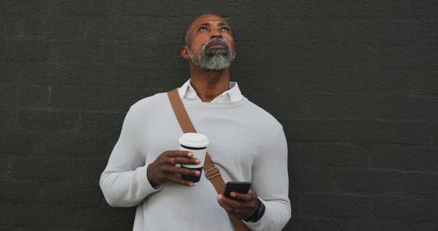 Bearded man in casual wear holding a coffee cup and smartphone, standing in urban environment and looking towards the sky. This versatile image can be used for marketing related to coffee shops, smartphones, or urban lifestyles. It is also useful for content around relaxation, contemplation, or modern life.