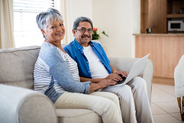 Senior couple sitting on sofa using laptop in living room. Perfect for themes on elderly lifestyle, retirement, technology adoption among seniors, and family togetherness.