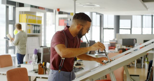 Young biracial man works on a project in a modern office. He focuses intently while a Caucasian man is busy in the background.