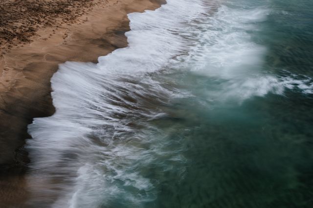 The waves softly washing onto a sandy beach, making intricate patterns with the seafoam. A notion of tranquil and untouched nature, perfect for outdoor and travel blogs, tourism advertisements, or backgrounds for presentations emphasizing peace and calmness.