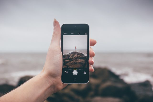 Close-up of a hand holding a smartphone, taking a photograph of a seaside landscape with rocky shore and cloudy sky. Ideal for themes of technology in nature, capturing moments, social media, and mobile photography.