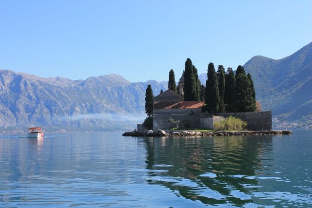 This image captures a remote island with a historic stone structure and tall cypress trees in a calm lake, framed by towering mountains under a clear blue sky. There is a boat visible on the left part of the image, enhancing the travel and adventure aspect. This scene is perfect for travel promotions, nature documentaries, Mediterranean tours, and pictorial exploration guides.