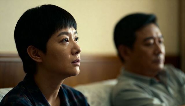 Asian woman sitting on bed gazing thoughtfully while partner sits blurred in background