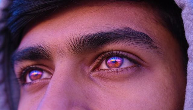 Neon-reflective iridescent eyes close-up of adult male in gray hood, moody cyberpunk gaze