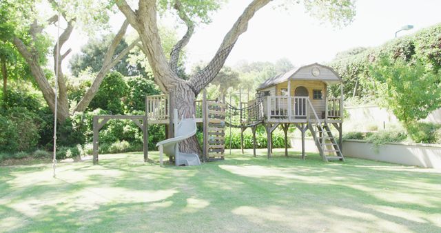 Treehouse with slide and wooden play area in bright, sunny backyard. Large tree providing shade and integrated into play structure. Green lawn and lush garden surround playground. Perfect for illustrating family fun, outdoor play, and summer activities. Ideal for parenting magazines, landscaping advertisements, and children's lifestyle content.