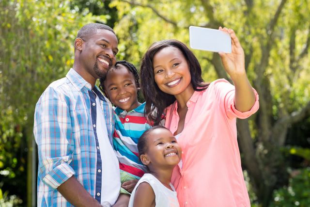 This image features a happy African American family taking a selfie outdoors in a park. The family, consisting of parents and children, is smiling and enjoying a moment of togetherness in nature. Perfect for use in advertisements, social media posts, lifestyle blogs, and websites promoting family values, outdoor activities, and technology.