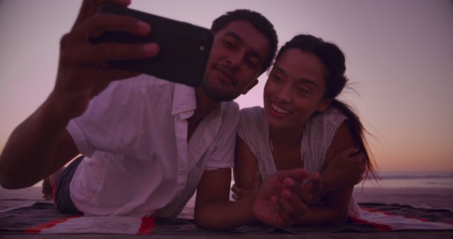 Couple enjoying a romantic evening. They are lying down on a beach blanket, capturing a selfie at sunset. Suitable for love, travel, vacation, summer, romance, and social media posts.