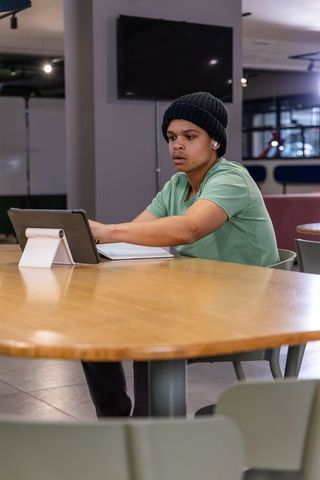 Young man working on tablet in coworking lounge wearing beanie and earbuds
