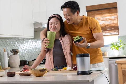 Asian couple preparing and tasting green smoothie at modern kitchen island with blender