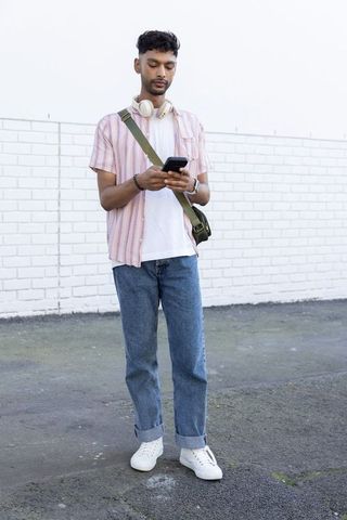 Indian young man standing by white brick wall using smartphone with headphones