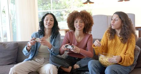 Diverse women playing video games on couch, laughing and snacking in bright living room