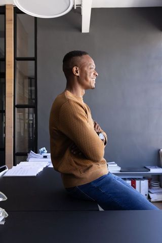 African american man sitting on desk edge smiling confidently in minimalist modern office