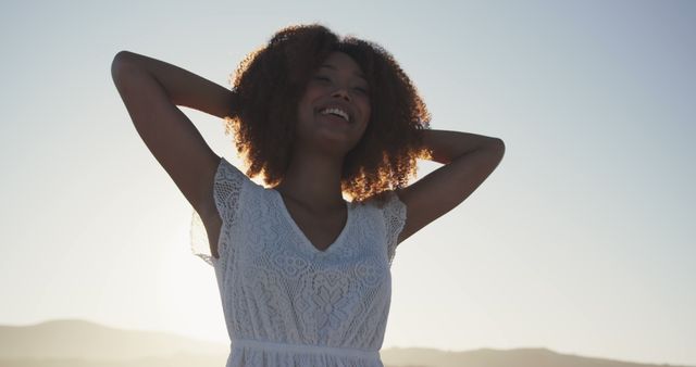 African American woman stands outdoors at sunset, wearing a white lace dress and smiling blissfully with arms raised. The image is perfect for themes related to happiness, freedom, nature, and carefree living. Can be used in marketing materials, lifestyle blogs, mental health and wellness articles, and advertisements promoting positive emotions and outdoor activities.