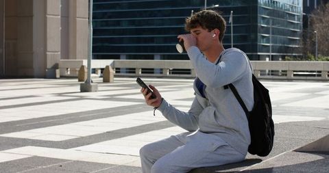 Young man sipping coffee and checking smartphone while sitting on ledge in urban plaza