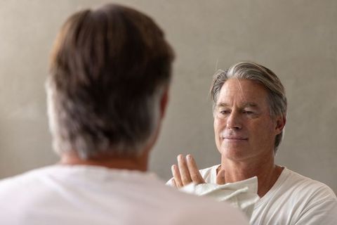 Mature man grooming in bathroom mirror holding towel, calm daily self-care routine for men