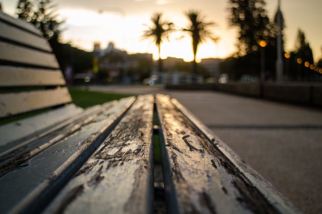 Worn wooden bench photographed at sunset in a city park with blurred background; suitable for themes of relaxation, urban lifestyle, peaceful moments, evening leisure, and the beauty of nature in urban settings.
