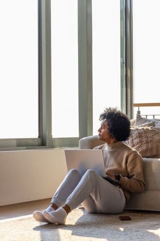 African american woman sitting on rug with laptop looking out window, remote work focus
